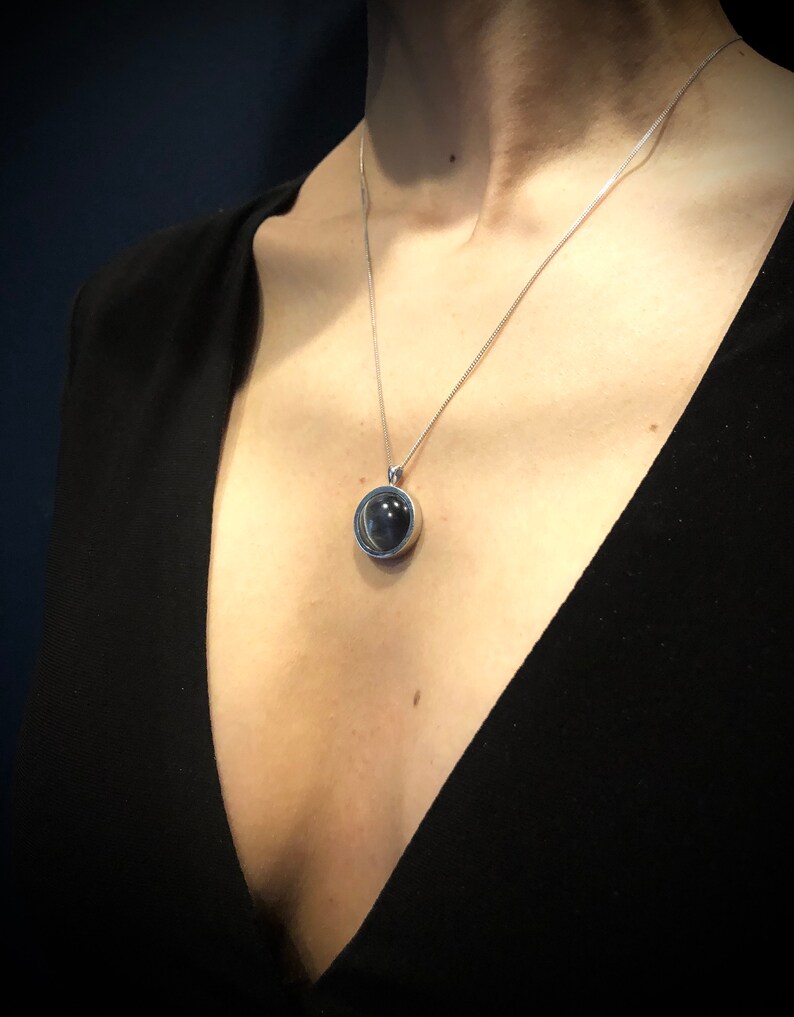 Op de afbeelding: Een zilveren ketting met een ronde hanger met een donkergrijze steen in een heldere glazen bol.