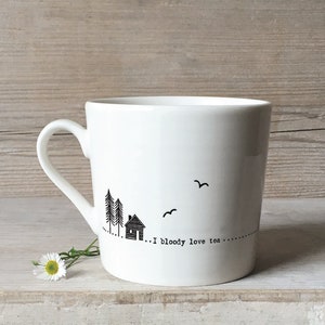 May include: White ceramic mug with a black illustration of a house, trees, and birds. The mug has a black text that reads "... I bloody love tea ..."