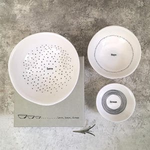 May include: A set of three white ceramic bowls with black text and designs. The largest bowl has the word "Love" and a dotted pattern. The other bowls say "Hope" and "Dream". The bowls are on a grey box that says "Love, hope, dream".