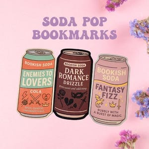 May include: Three colorful soda can-shaped bookmarks with different designs and text. The first bookmark is orange and green with the text "Bookish Soda Enemies to Lovers Cola Spicy, Chaotic, Unforgettable". The second bookmark is brown with the text "Bookish Soda Dark Romance Drizzle Bittersweet and addicting". The third bookmark is pink with the text "Bookish Soda Fantasy Fizz Bubbly with a burst of magic."