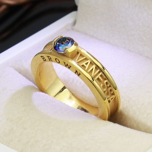 May include: A gold ring with the names "Vanessa" and "Brown" engraved on the band. The ring has a blue gemstone in the center.
