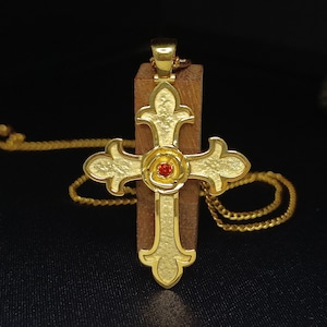 May include: A gold-tone cross pendant with a red gemstone in the center of a rose design. The pendant is on a gold chain.