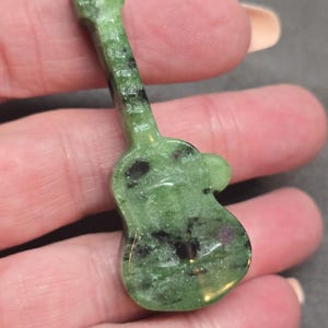 May include: A small, green gemstone guitar pendant with black inclusions. The guitar-shaped pendant is held in a hand, showcasing its polished surface and detailed carving. The gemstone has a translucent quality.