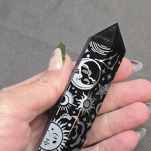 May include: Black crystal obelisk with etched celestial designs. The polished stone features a sun, moon, and star pattern in silver. The obelisk is held in a hand, showcasing its detailed artwork and pointed top.