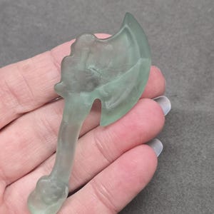 May include: A translucent, light green axe-shaped carving. The miniature axe features a detailed handle and blade, held in a hand against a gray background. The carving is likely made of glass or a similar material.