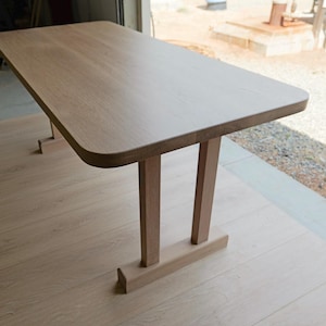 May include: A light wood table with a rounded edge and two simple, rectangular legs. The table is made of solid wood and has a natural finish.