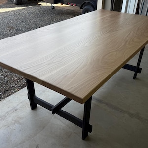 May include: A large wooden table with a natural wood finish and a black metal base. The table has a rectangular top and a simple, modern design.