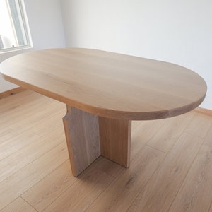 May include: A light wood oval table with a simple, modern design. The table has a single, wide base that supports the oval tabletop.