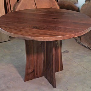 May include: A round, solid wood table with a dark brown finish. The tabletop is supported by a pedestal base with a unique, flared design. The table is approximately 36 inches in diameter.