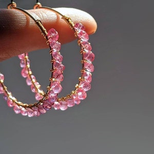 Op de afbeelding: Een paar gouden creolen met roze edelsteen accenten. De oorbellen zijn gemaakt van draad en hebben een delicate, vrouwelijke uitstraling.