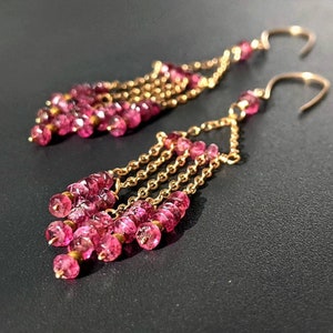 Könnte beinhalten: Ein Paar goldene Ohrringe mit rosafarbenen Edelsteinperlen, die an einer Kette hängen. Die Ohrringe sind im Hänge-Stil und haben eine zarte Optik.