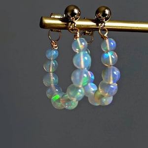 May include: A pair of gold hoop earrings with iridescent opal beads. The earrings are hanging from a gold stand.