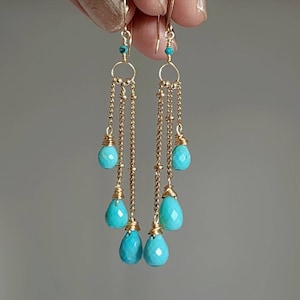 Puede incluir: Pendientes de aro dorados con piedras turquesa. Los pendientes tienen múltiples cadenas de oro con piedras turquesa colgando de cada cadena.