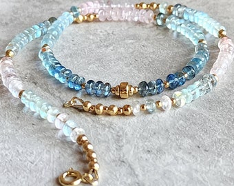 May include: A delicate necklace featuring a strand of small, light blue, and pink gemstones interspersed with gold-colored beads. The necklace has a gold clasp and is displayed on a light gray surface.