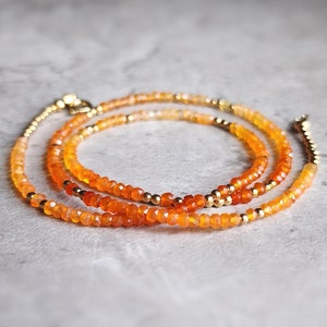 May include: A layered necklace featuring strands of orange and gold beads. The necklace has a delicate design with small, faceted orange beads and interspersed gold-colored beads. The clasp and connecting elements are also gold-toned.