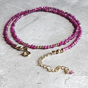 Pu&ograve; includere: Una delicata collana con piccole pietre preziose rosa intenso sfaccettate. Perline color oro e una chiusura e catena di estensione color oro aggiungono contrasto. La collana &egrave; esposta su una superficie strutturata grigio chiaro.