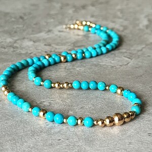 May include: A turquoise and gold beaded necklace. The necklace is made of small, round turquoise beads and gold beads. The necklace is on a gray surface.