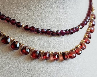 Könnte beinhalten: Eine zweireihige Halskette mit einer goldenen Kette und roten Edelsteinperlen. Die untere Reihe hat größere, facettierte, tropfenförmige Perlen.