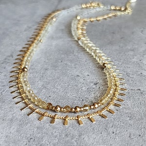 Könnte beinhalten: Eine goldene Halskette mit zwei Reihen Perlen. Die obere Reihe besteht aus klaren Perlen und die untere Reihe aus hellgelben Perlen. Die Halskette hat eine goldene Kette mit kleinen, spitzen goldenen Anhängern.