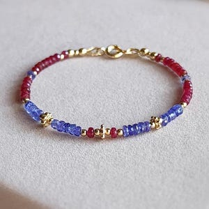 May include: A delicate bracelet featuring alternating segments of small, faceted red and blue gemstone beads. The design is accented with tiny gold-toned metal beads and small, flower-shaped gold-toned spacers, finished with a gold-toned circular clasp.