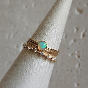 May include: Three gold rings are displayed on a white cone. One ring features a round opal stone, while another has a row of small, clear stones. The rings are stacked, showcasing a minimalist jewellery design.