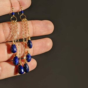 May include: Gold-toned dangle earrings featuring faceted, deep blue lapis lazuli teardrop beads. The earrings have delicate gold chains and small blue beads at the top. The design is elegant and eye-catching, perfect for adding a touch of color.