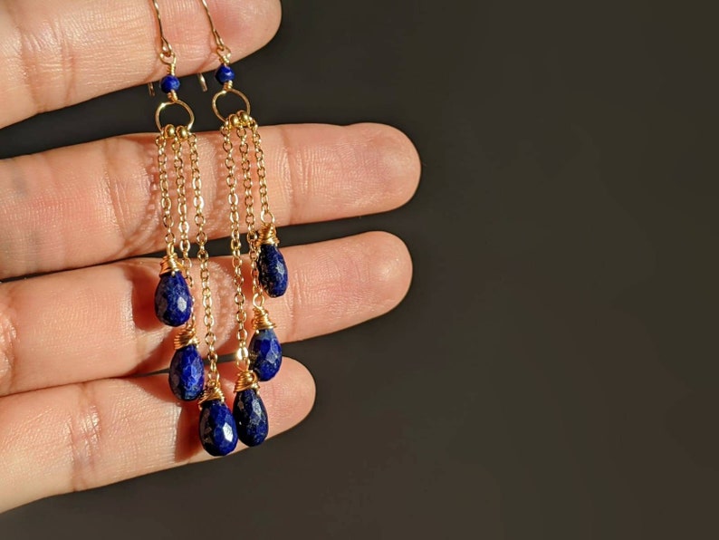May include: Gold-toned dangle earrings featuring faceted, deep blue lapis lazuli teardrop beads. The earrings have delicate gold chains and small blue beads at the top. The design is elegant and eye-catching, perfect for adding a touch of color.