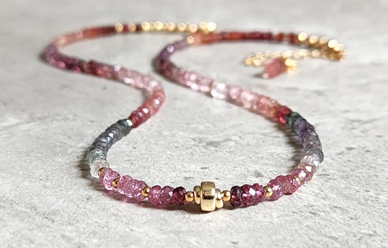 May include: A necklace with a gradient of pink, purple, and red gemstones. The necklace has a gold clasp and small gold beads. The gemstones transition from darker to lighter shades, creating a visually appealing accessory.