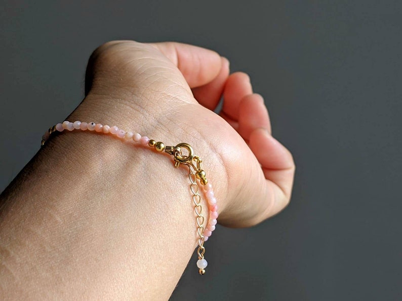 May include: A delicate bracelet made with small pink opal beads and gold accents. The bracelet has a lobster clasp closure and an adjustable chain.