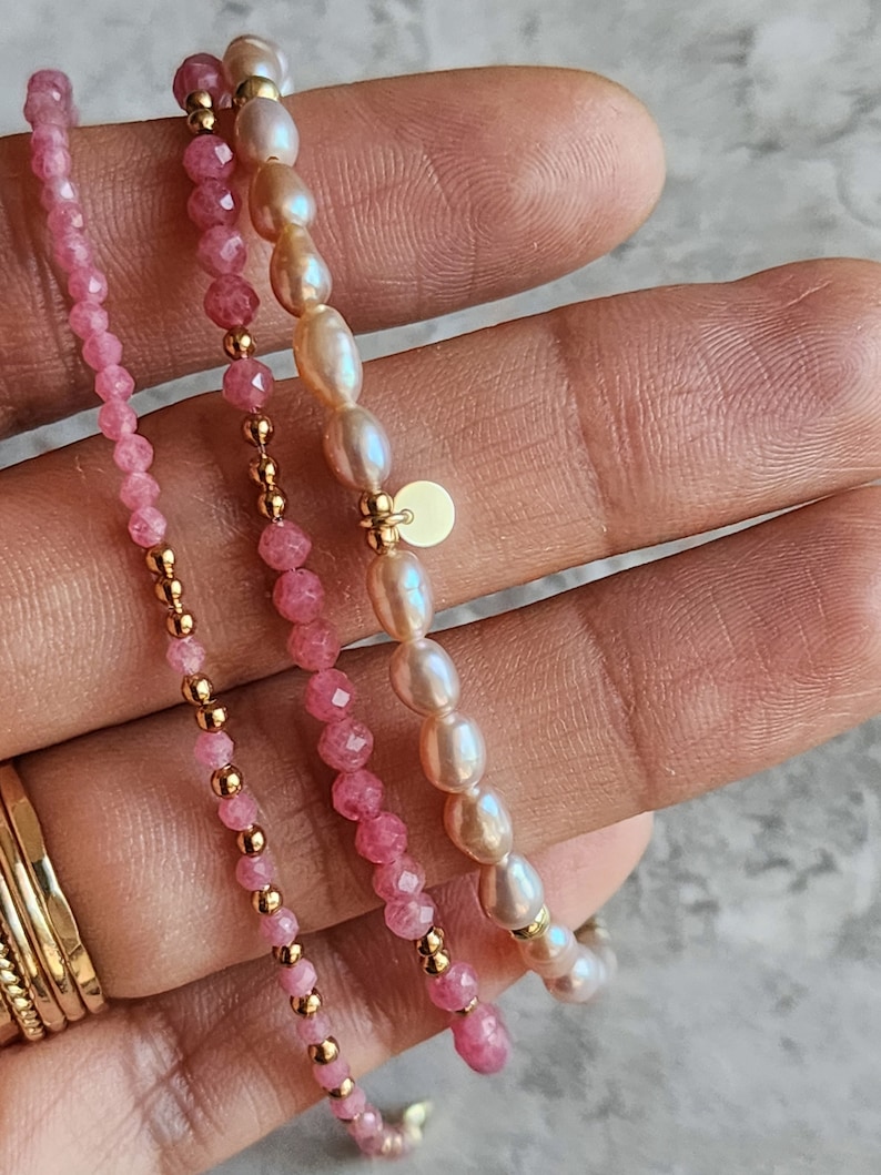 Puede incluir: Tres pulseras delicadas. Una est&aacute; ensartada con peque&ntilde;as cuentas rosas facetadas y detalles dorados. Otra presenta perlas rosas y cuentas doradas. La tercera pulsera est&aacute; hecha de perlas rosas y detalles dorados. Las pulseras se muestran sobre un fondo claro.