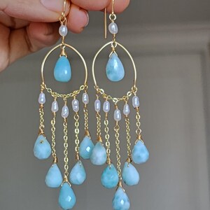 May include: Gold-toned chandelier earrings featuring teardrop-shaped, light blue gemstones and small white pearls. The earrings have a circular design with dangling chains and gemstones, creating a layered effect. The earrings are held up by a hand.