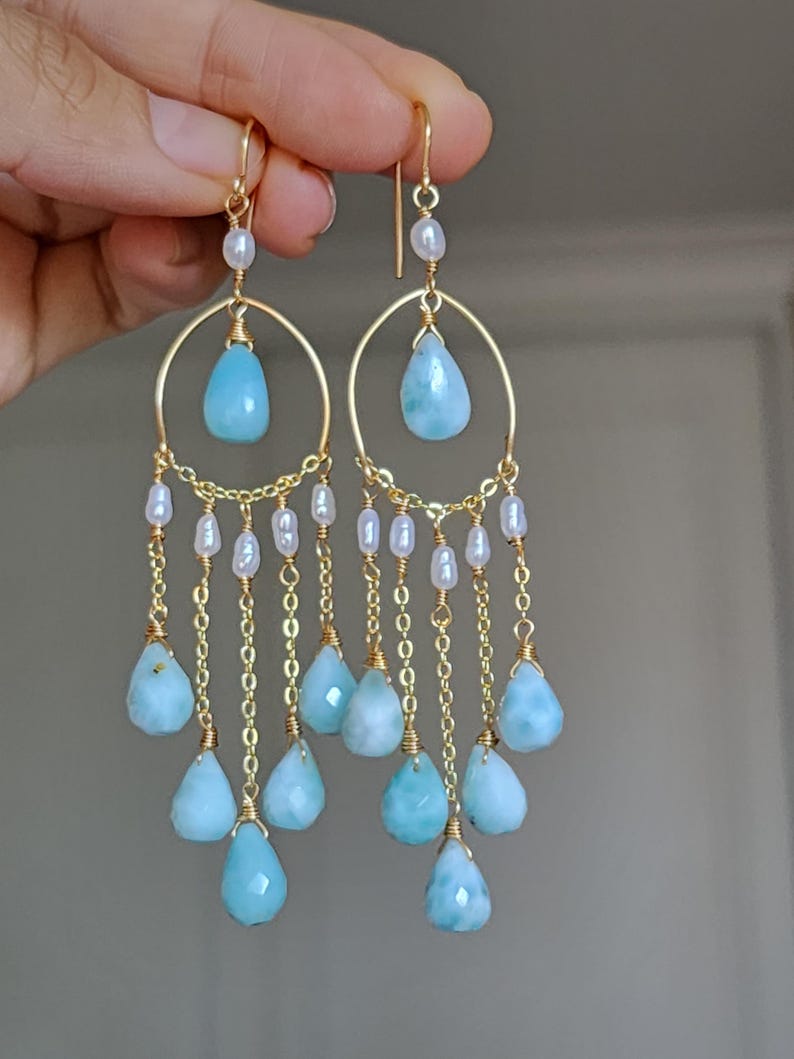 May include: Gold-toned chandelier earrings featuring teardrop-shaped, light blue gemstones and small white pearls. The earrings have a circular design with dangling chains and gemstones, creating a layered effect. The earrings are held up by a hand.