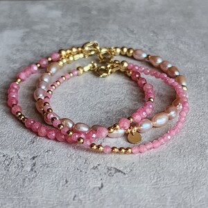 Puede incluir: Tres delicadas pulseras con piedras preciosas rosas, perlas y cuentas doradas. Las pulseras est&aacute;n superpuestas, mostrando una variedad de texturas y tonos de rosa. Cada pulsera tiene un cierre dorado.