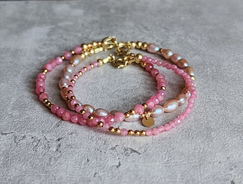 Puede incluir: Tres delicadas pulseras con piedras preciosas rosas, perlas y cuentas doradas. Las pulseras est&aacute;n superpuestas, mostrando una variedad de texturas y tonos de rosa. Cada pulsera tiene un cierre dorado.