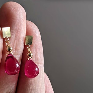 May include: A pair of gold earrings with teardrop-shaped red gemstones hanging from square gold charms.