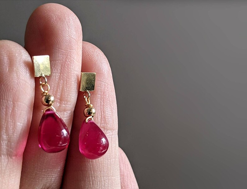 May include: A pair of gold earrings with teardrop-shaped red gemstones hanging from square gold charms.