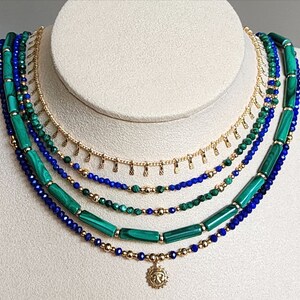 May include: A layered necklace with a gold chain and green and blue beads. The necklace has a small sun pendant at the bottom.