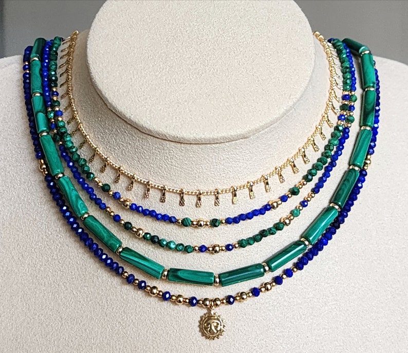 May include: A layered necklace with a gold chain and green and blue beads. The necklace has a small sun pendant at the bottom.