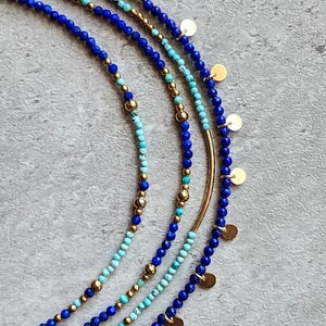 May include: A set of four layered necklaces featuring blue and turquoise gemstone beads and gold accents. The necklaces are adorned with small gold discs.