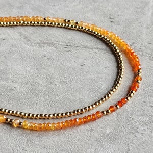May include: Two layered necklace with gold-colored beads and orange and yellow gemstones. The gemstones transition in color from yellow to orange to red. The necklace is on a gray surface.