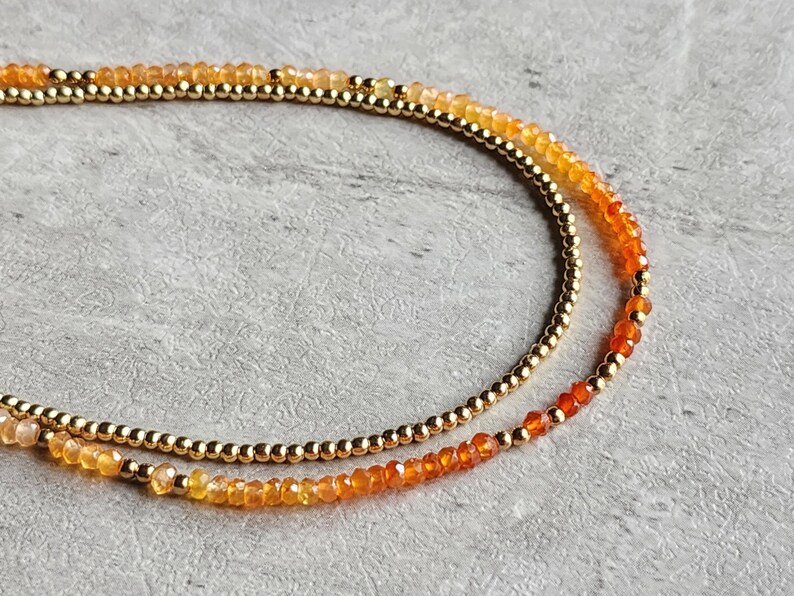 May include: Two layered necklace with gold-colored beads and orange and yellow gemstones. The gemstones transition in color from yellow to orange to red. The necklace is on a gray surface.