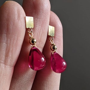 May include: A pair of gold earrings with teardrop-shaped red glass stones hanging from square gold charms.