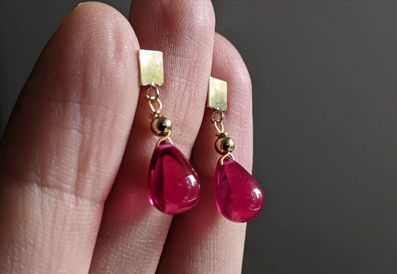 May include: A pair of gold earrings with teardrop-shaped red glass stones hanging from square gold charms.