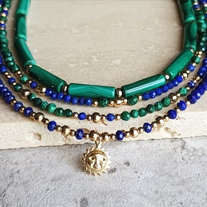 May include: A layered necklace with blue lapis lazuli and green malachite beads, accented with gold beads and a gold sun charm.