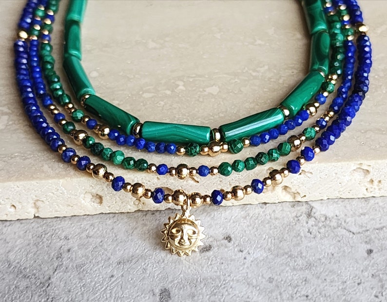 May include: A layered necklace with blue lapis lazuli and green malachite beads, accented with gold beads and a gold sun charm.