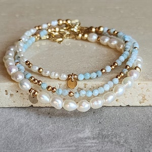 May include: Three layered bracelets with pearls, light blue beads, and gold-colored accents. Each bracelet features a different design, with a mix of round and irregular-shaped pearls. Small gold-colored charms dangle from the bracelets.