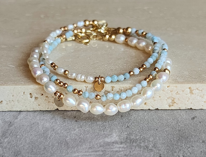 May include: Three layered bracelets with pearls, light blue beads, and gold-colored accents. Each bracelet features a different design, with a mix of round and irregular-shaped pearls. Small gold-colored charms dangle from the bracelets.
