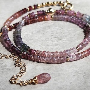 May include: A multi-coloured gemstone necklace with a gold chain and clasp. The necklace features a gradient of pink, purple, and blue gemstones. A small, teardrop-shaped pendant hangs at the end. The gemstones are small and round, creating a subtle, elegant look.