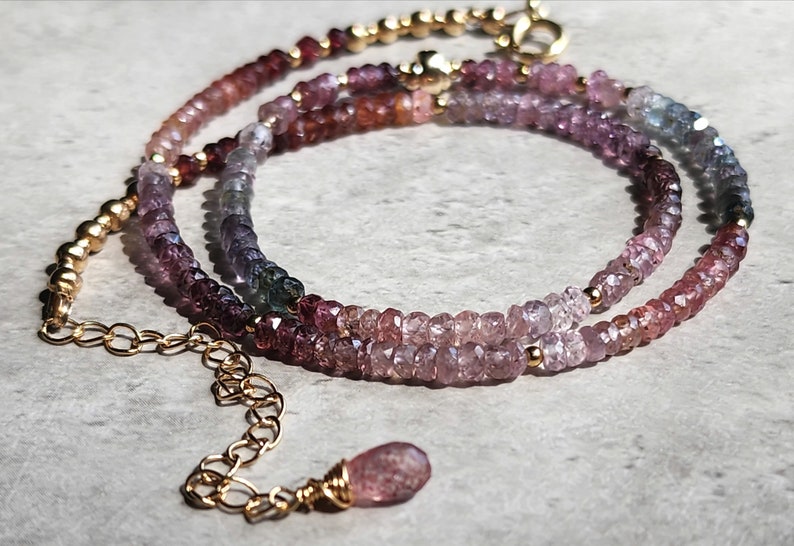 May include: A multi-coloured gemstone necklace with a gold chain and clasp. The necklace features a gradient of pink, purple, and blue gemstones. A small, teardrop-shaped pendant hangs at the end. The gemstones are small and round, creating a subtle, elegant look.
