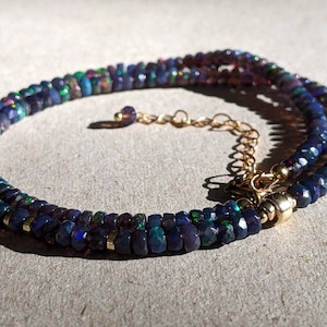 Op de afbeelding: Een ketting met gefacetteerde donkerblauwe en zwarte opaal kralen met iriserende groene en rode flitsen. De streng is geaccentueerd met kleine goudkleurige metalen tussenstukjes en bevat een goudkleurige metalen verlengketting met karabijnsluiting.